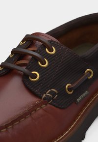 Καφέ δερμάτινο παπούτσι τύπου boat με σκούρα καφέ κορδόνια, χρυσές πόρπες και ραφτά διακοσμητικά στοιχεία, εμφανίζοντας την ετικέτα της μάρκας Barbour στην πλευρά.