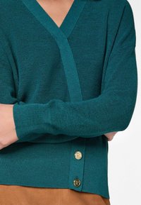 Teal gebreide trui met een V-halsontwerp, geribbeld textuur en gouden knopen aan de zoom. Beschikt over lange mouwen en een losse pasvorm.