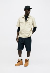 Άνδρας που φοράει μπεζ πουκάμισο με κοντά μανίκια πάνω από μαύρα μακριά μανίκια, σκούρα σορτς μέχρι το γόνατο, ταμπά εργασιακά μποτάκια, άσπρες κάλτσες και μαύρο καπέλο, όρθιος.