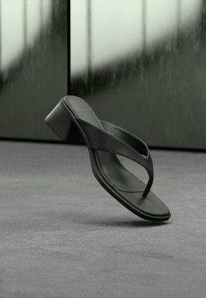 Sandalo infradito nero con tacco e ampi cinturini che fluttua sopra un tappeto grigio e pannelli di sfondo scuri e sfocati.