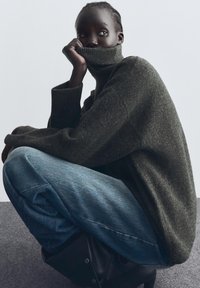 Dark green oversized sweater with a tall collar, worn with loose blue denim jeans. The fabric appears textured and soft.