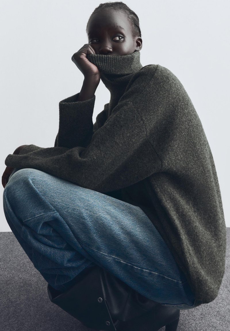 Dark green oversized sweater with a tall collar, worn with loose blue denim jeans. The fabric appears textured and soft.