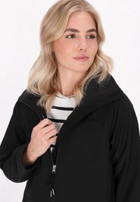 Juodas minkštas striukė su užtrauktuku, gobtuvu ir išsiuvinėtu skėčio logotipu. Dėvima ant baltos ir juodos juostuotos marškinėlių, turinčių lygų paviršių.