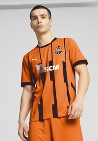 Camiseta de fútbol naranja con rayas verticales negras, de mangas cortas, cuello redondo y el logo de Puma. Incluye el escudo del club y un gráfico de patrocinador.