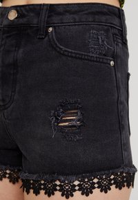 Nära bild av svarta denimshorts med slitna hål och svart blommig spetskant som bärs på övre låret.