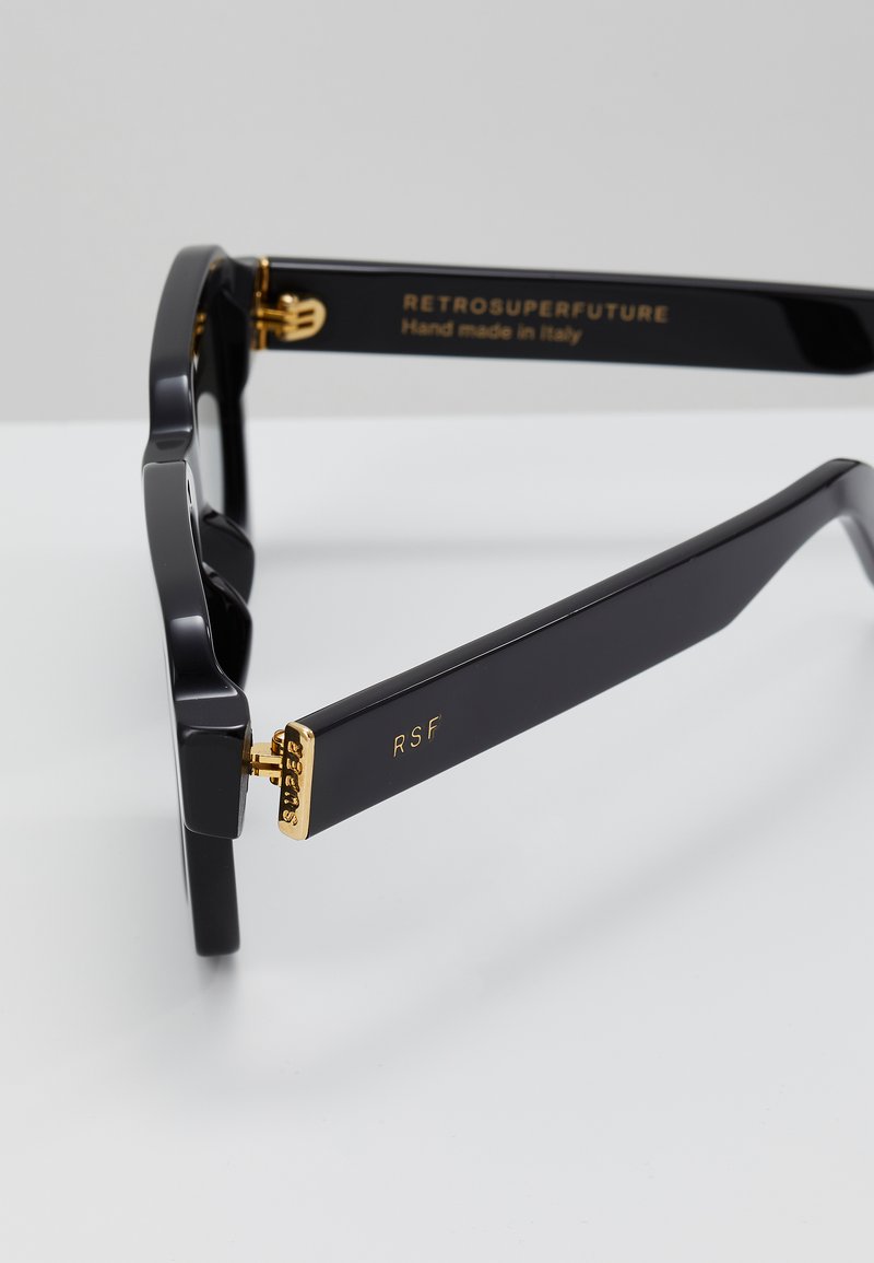 Lunettes de soleil en acétate noir avec une forme rectangulaire audacieuse, présentant des accents dorés sur les charnières et le logo. Finition texturée sur la monture.