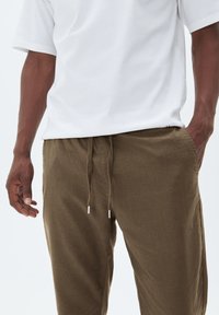 Weißes Kurzarm-T-Shirt, kombiniert mit olivgrünen Jogginghosen mit Kordelzug aus strukturiertem Stoff und silbernen Metallspitzen an den Kordeln.