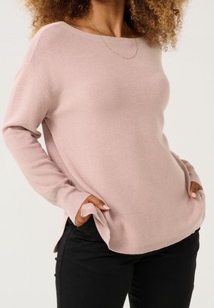 Femme aux cheveux bouclés portant un pull côtelé rose clair et un pantalon noir, les mains dans les poches avant, sur un fond uni.