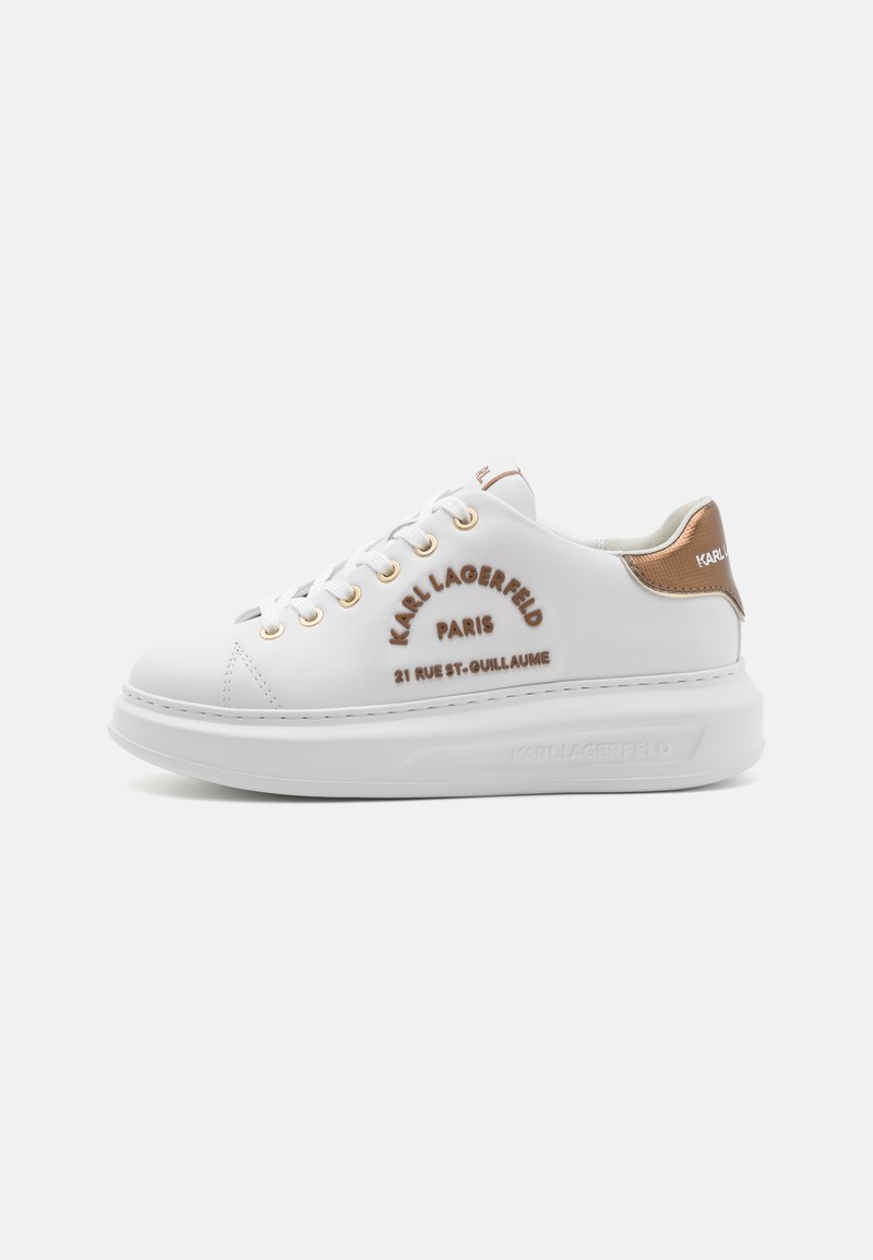 White sneakers with branding in brown, gold eyelets, and a textured brown heel accent. Features a streamlined design with a thick sole.