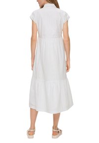 Witte katoenen jurk met korte capmouwen, een kraag, een gelaagde rok en details van oogjes borduursel. Van achteren getoond met platte sandalen.