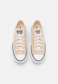 Sneakers in tela beige chiaro con lacci bianchi, punta in gomma e accento nero sulla suola. Presentano il logo "Converse All Star" sulla linguetta.