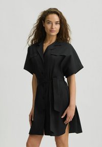 Abito camicia nero con maniche corte, chiusura a bottoni davanti, due tasche sul petto, cintura in vita e spacchi laterali. Realizzato in tessuto leggero con una consistenza liscia.