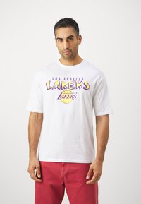 Bijela pamučna majica s šarenim grafičkim logotipom "Los Angeles Lakers" u ljubičastoj i žutoj boji. Okrugli izrez, kratki rukavi, opušteni kroj.