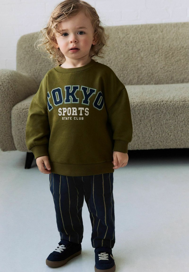 Oliwkowy, oversize'owy sweatshirt z grafiką "TOKYO SPORTS", w zestawie z granatowymi spodniami w paski i czarnymi trampkami z białymi sznurowadłami.