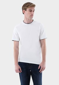 Weißes Kurzarm-T-Shirt mit rundem Halsausschnitt und kontrastierendem schwarzen Besatz. Der Stoff wirkt glatt und leicht tailliert, kombiniert mit blauen Jeans.
