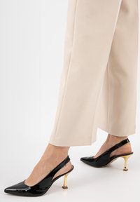 D�écolleté in vernice nera con punta affilata e tacco a blocco dorato, abbinate a pantaloni beige a gamba larga.