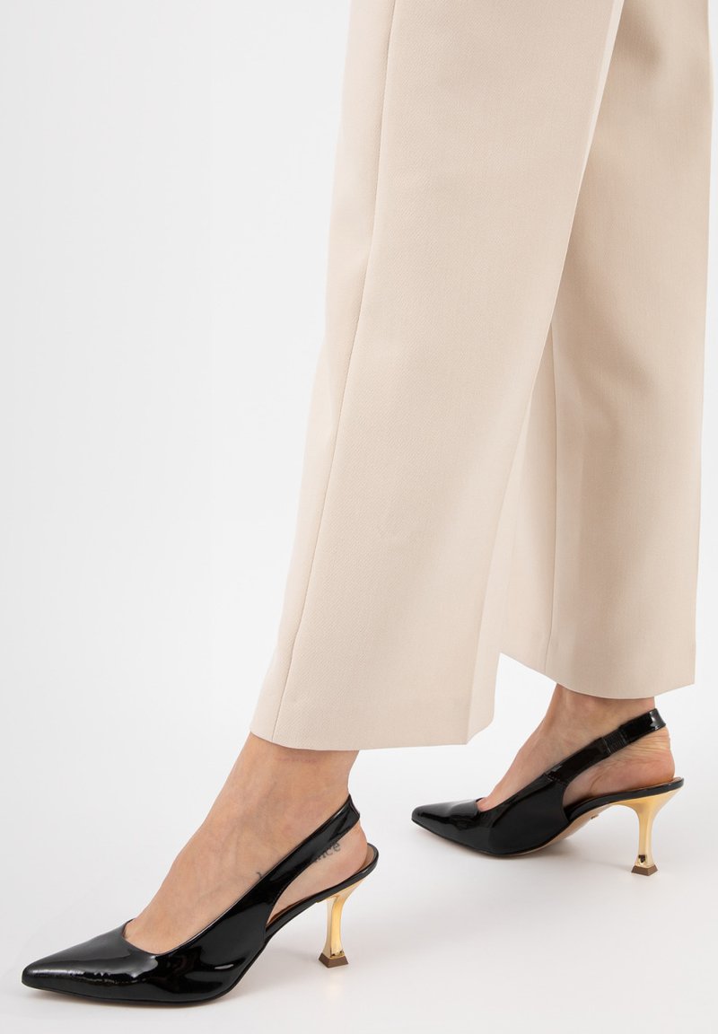 Décolleté in vernice nera con punta affilata e tacco a blocco dorato, abbinate a pantaloni beige a gamba larga.