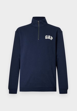 Temno modra pulover s pol dolgimi rokavi in polovičnim zadrgo na ovratniku ter belim logotipom »GAP« na levi strani prsnega koša.