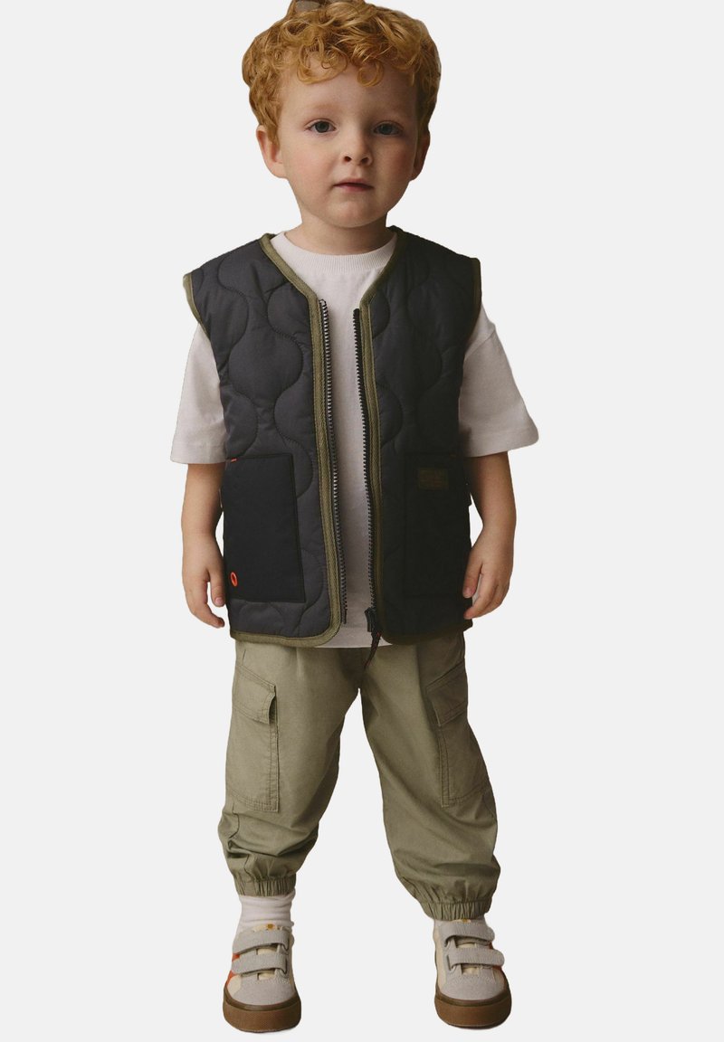 Vestă neagră matlasată cu margini olive, două buzunare frontale, purtată peste un tricou alb, asortată cu pantaloni cargo olive și pantofi bej.