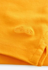 Tela de algodón naranja con un sutil logo bordado. Textura suave con bordes cosidos de forma ordenada y un dobladillo doblado.