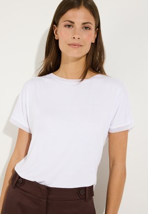 Jonge vrouw met steil bruin haar, draag een simpel wit T-shirt met korte mouwen en donkerbruine broek met hoge taille tegen een lichte achtergrond.