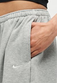 Χέρι που ακουμπά στην μπροστινή τσέπη ανοιχτόχρωμου γκρι παντελονιού Nike με ελαστική μέση, εμφανίζοντας μέρος της μαύρης άνω άκρης.