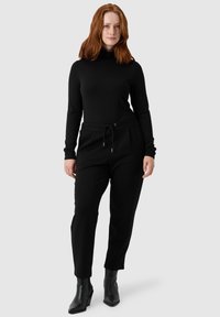 Maglione a maniche lunghe nero con collo alto abbinato a pantaloni neri tapered con elastico in vita e cordino. Indossato con stivali neri con tacco.