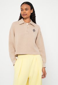 Pólo bege de mangas compridas com colarinho, três botões e um logótipo de diamante preto. Combinado com calças de cintura alta amarelo claro.