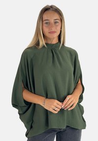 Grøn, løstsiddende top med høj hals, tekstureret stof og sideslidser. Modellen bærer den med mørke jeans og enkelt sølvsmykker.