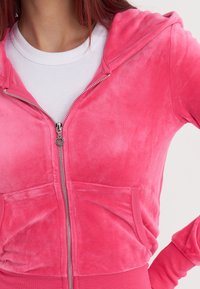 Mujer vestida con una sudadera de felpa rosa brillante con cremallera y tirador en forma de corazón, sobre una camiseta blanca de canalé, con la mano en la cadera.