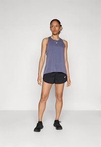 Fioletowy bezrękawnik sportowy z okrągłym dekoltem, w zestawieniu z czarnymi szortami. Model nosi czarne buty sportowe, stoi na jednolitym tle.