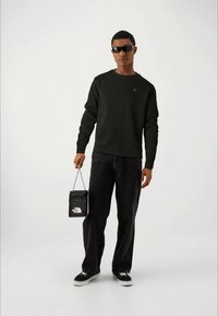 Zwarte crewneck sweatshirt, zwarte jeans, zwarte sneakers, houdt een kleine zwarte tas met een wit logo vast. Strakke, casual pasvorm overal.