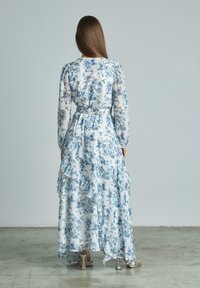 Florales Maxikleid in Weiß und Blau, mit langen Ärmeln, Rüschenschichten und tailliertem Bund. Hergestellt aus leichtem, durchsichtigem Stoff.