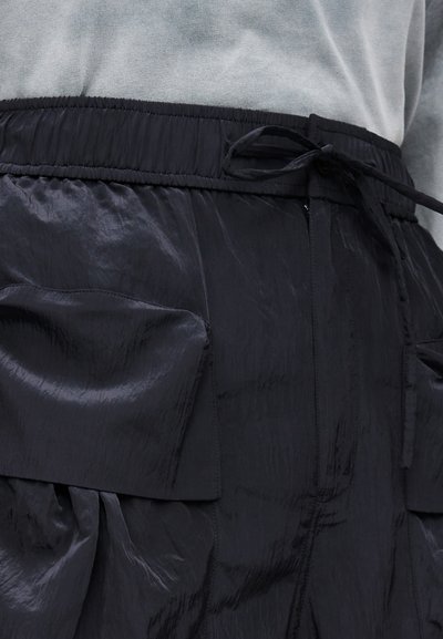 Μαύρο γυαλιστερό παντελόνι με μεγάλες μπροστινές τσέπες και δεμένη ελαστική μέση, φορεμένο με γκρι πουκάμισο.