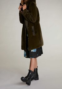 Manteau en fausse fourrure verte avec capuche, fermeture éclair à l'avant et poches latérales, associé à une robe noire à fleurs et des bottes de combat noires à lacets.