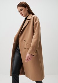 Cappotto cammello doppiopetto, realizzato in tessuto morbido con colletto a revers, dotato di due tasche frontali e bottoni color oro. Texture liscia.