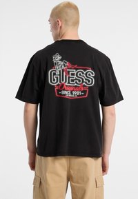 T-shirt nera a maniche corte con una grafica colorata "GUESS Originals" sul retro, caratterizzata da un design e un testo contornati.
