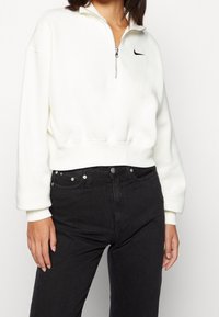 White zip-up sweatshirt with ribbed cuffs and hem, featuring a small black Nike logo on the chest, paired with high-waisted black jeans.