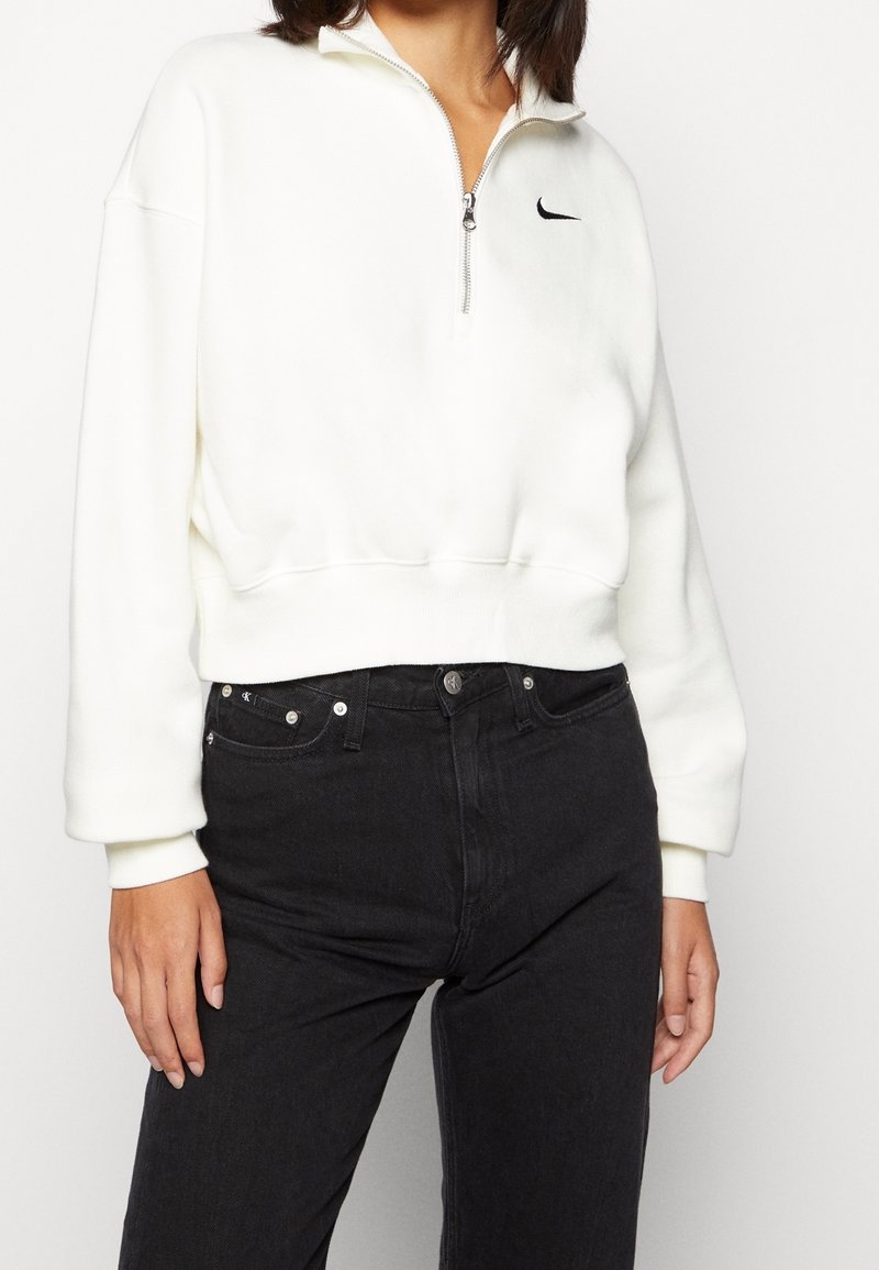 White zip-up sweatshirt with ribbed cuffs and hem, featuring a small black Nike logo on the chest, paired with high-waisted black jeans.