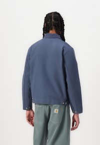 Jachetă din bumbac albastru cu guler, cusături vizibile și nasturi cu presiune. Pur ă peste pantaloni verzi, evidențiind o croială relaxată și un design casual.
