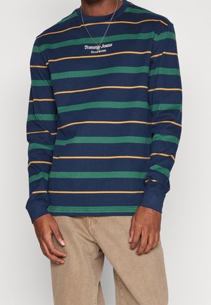 Homme portant une chemise à manches longues bleu marine avec des rayures horizontales vertes et moutarde, ainsi qu'un pantalon beige, se tenant les mains détendues.