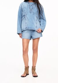 Felpa in denim azzurro con scollatura lace-up, maniche lunghe e due tasche frontali. Abbinata a pantaloni shorts in denim strappati e stivaletti beige.