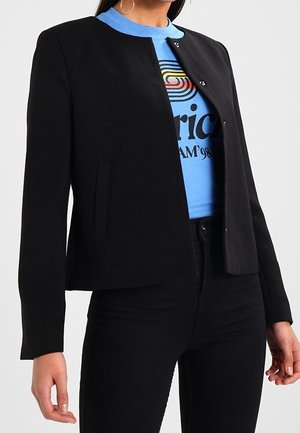 Femme portant une veste noire sans col boutonnée sur un T-shirt graphique bleu et un pantalon noir, montrant une vue partielle du torse.
