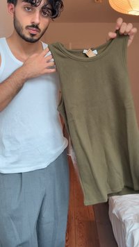 Oliwkowa bezrękawnikowa bluzka z prążkowanego materiału, z okrągłym dekoltem, pokazana z przymocowaną metką. Na tle jasnym z widoczną drewnianą podłogą.