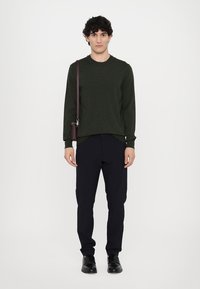 Dunkelgrüner Strickpullover, schwarze Hose und schwarze Schuhe. Das Model hält eine braune Umhängetasche. Einfaches Design, minimalistischer Stil.
