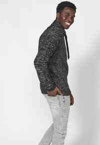 Pullover scuro con texture, colletto alto e cordoncino, abbinato a jeans slim-fit grigio chiaro. Vestibilità casual e comoda.