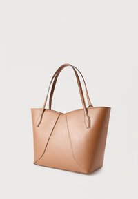 Τσάντα ώμου από δέρμα ταμπά με δύο λαβές και γωνιακές λεπτομέρειες ραφής σε απλό ανοιχτό φόντο.