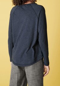Dunkelgraues Langarmshirt mit geribbelter Textur, abgerundetem Saum und Raglanärmeln, kombiniert mit karierten, weit geschnittenen Hosen. Der Hintergrund ist gelb.