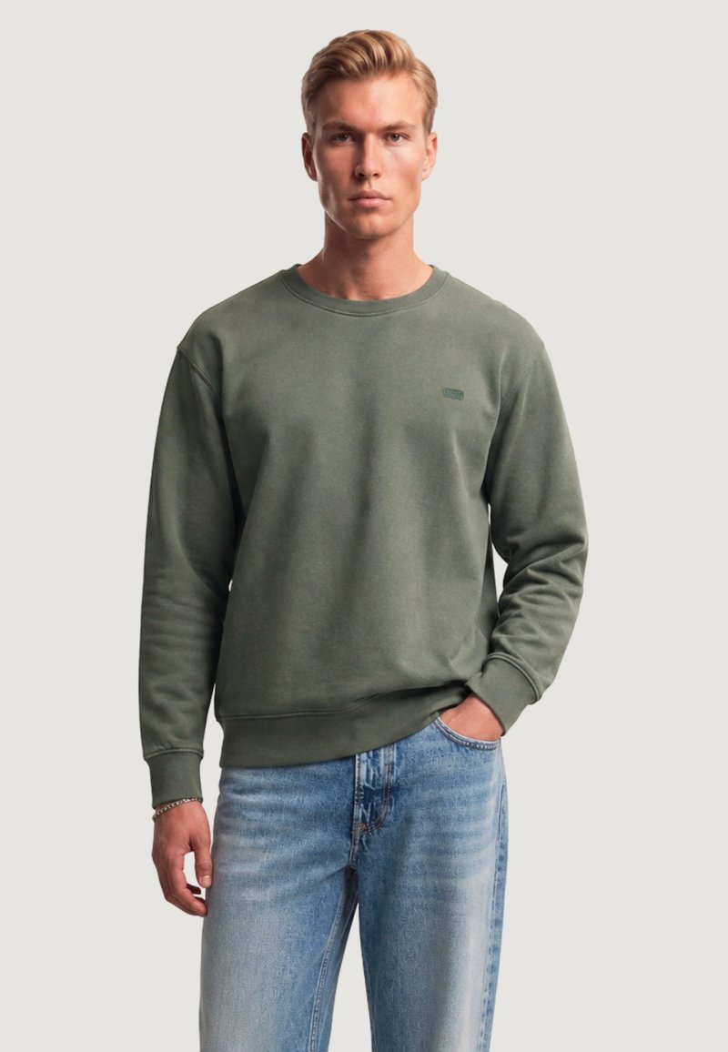 Homme aux cheveux blonds portant un sweat-shirt vert olive et un jean bleu clair, debout avec une main dans la poche devant un fond uni.