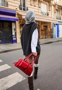 Červená lakovaná kožená kabelka s dvěma krátkými uchy, disponující několika kapsami, drží osoba oblečená v šedých pletených vestách a hnědých vysokých kozačkách.